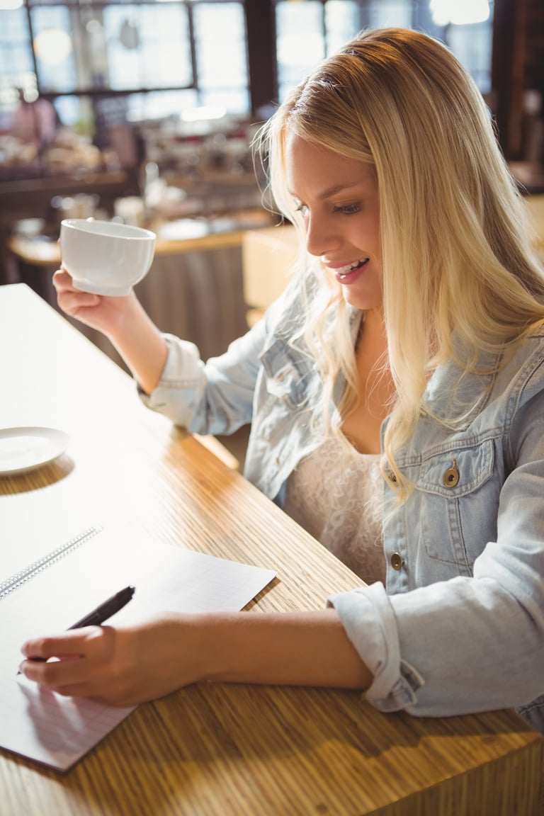 Smiling blonde drinking coffee and writing on sheet of paper